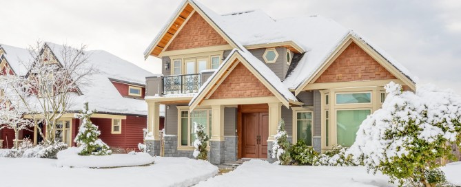 Grey and brown house with a wood front door and snow covering the yard and shrubbery.