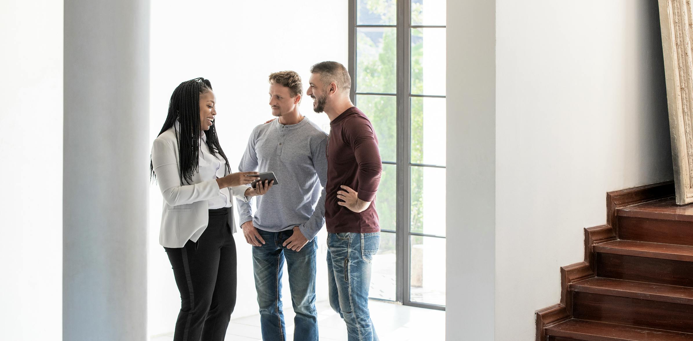 A real estate agent gives a house tour to prospective homebuyers. They are standing in a room with windows behind them.