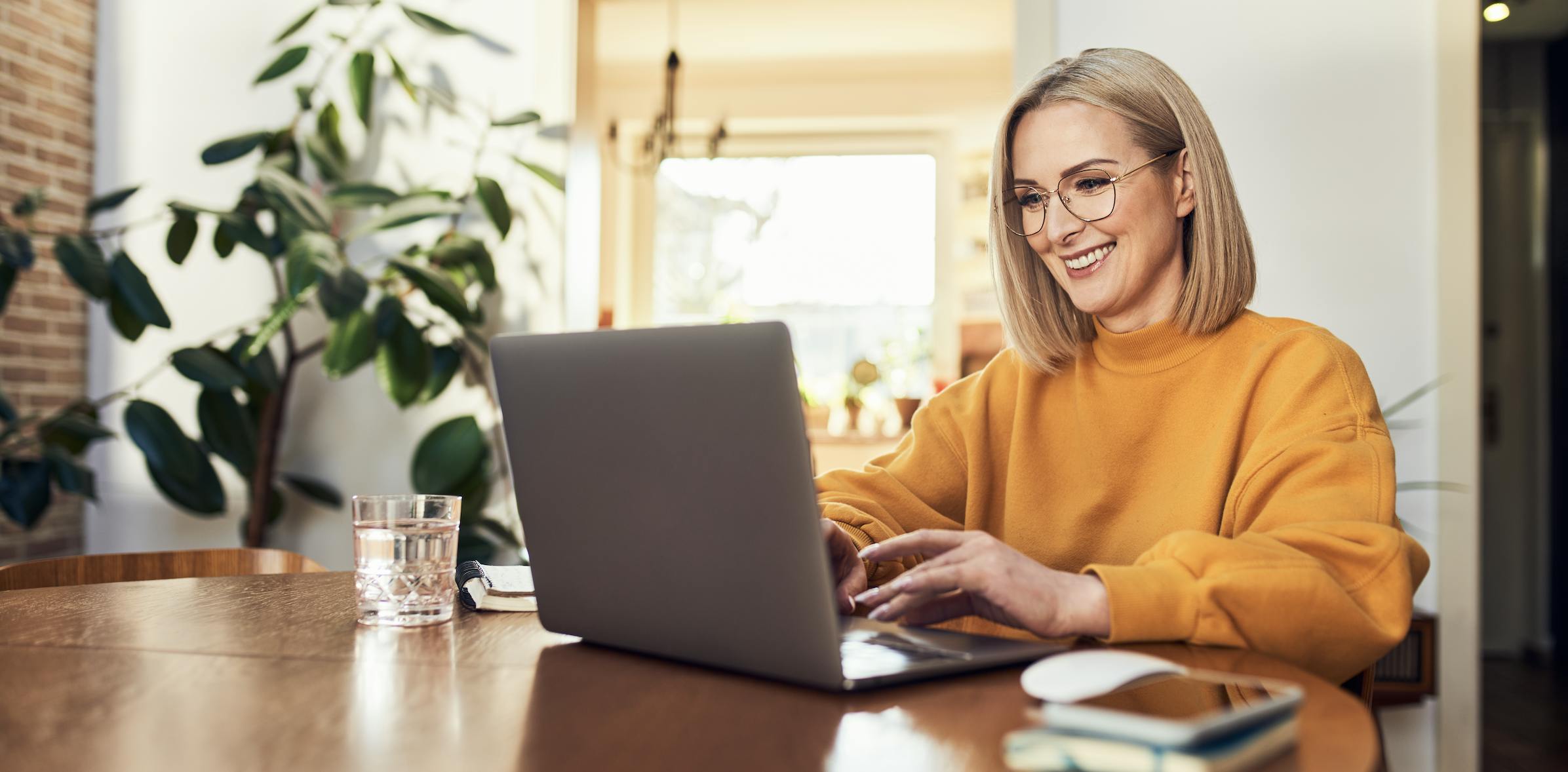 A woman sits at a table, wearing a yellow sweatshirt, and works on her laptop computer.